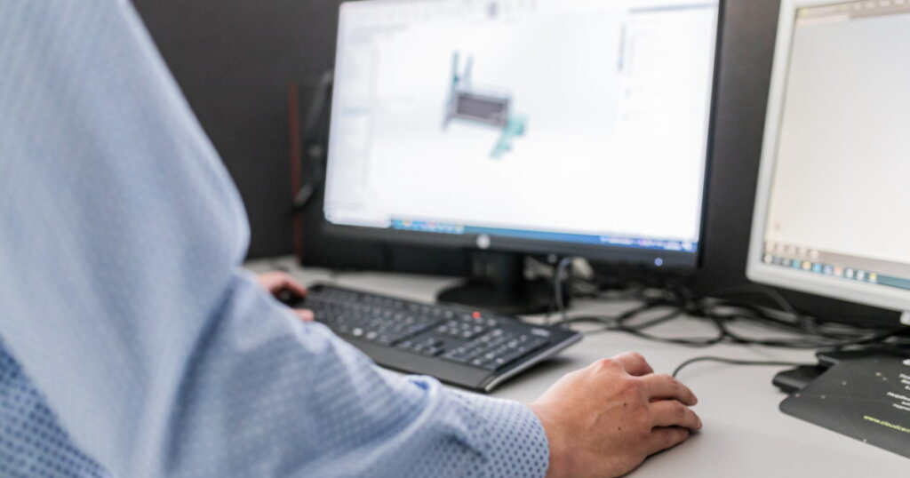 Henkilön käsi hiirellä, taustalla näppäimistäö ja kaksi näyttö, joilla näkyy suunnitelmia.